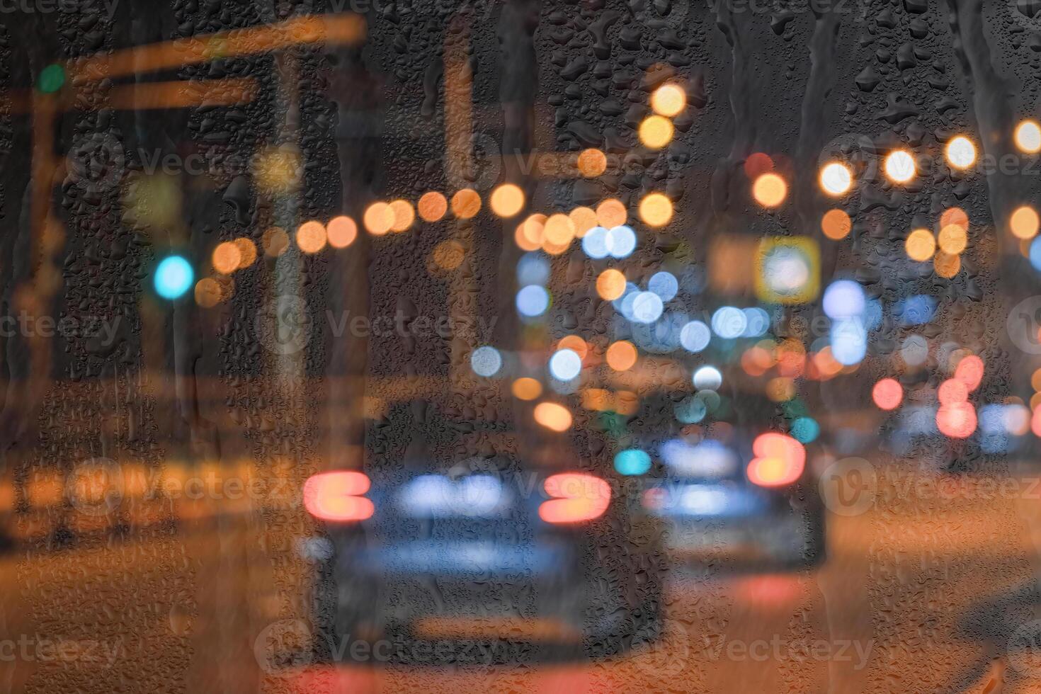 Blurry moving cars with headlights and lanterns reflecting on the wet asphalt in the night city behind rain-covered window. photo