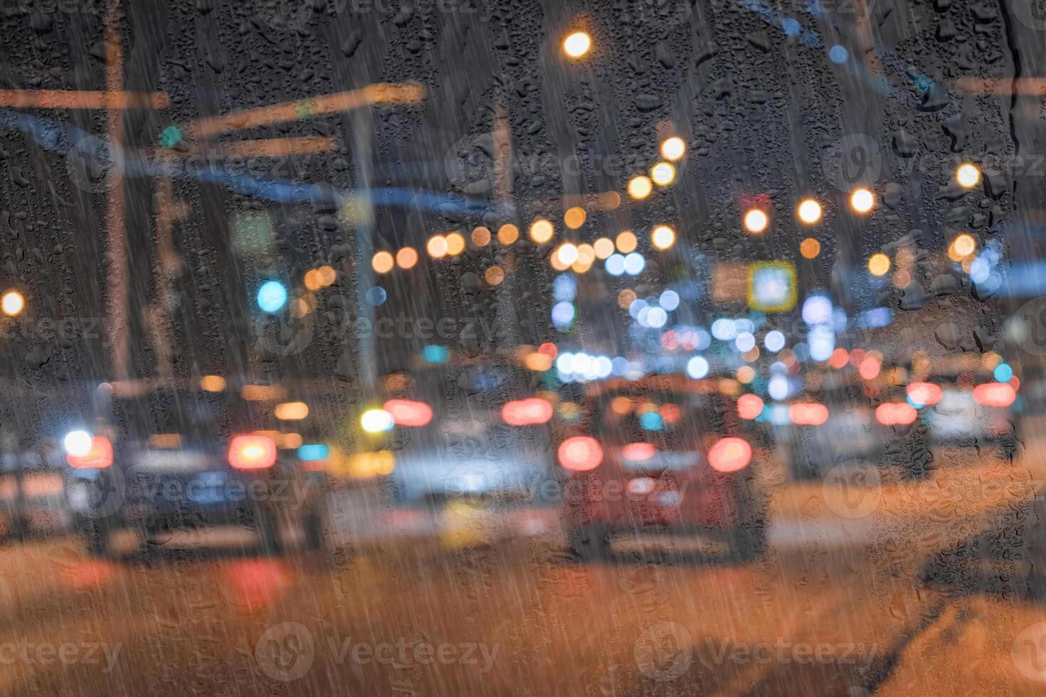 Blurry moving cars with headlights and lanterns reflecting on the wet asphalt in the night city behind rain-covered window. photo
