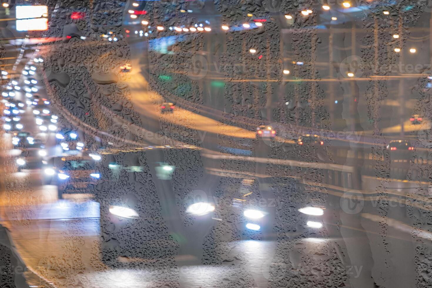 Blurry moving cars with headlights and lanterns reflecting on the wet asphalt in the night city behind rain-covered window. photo