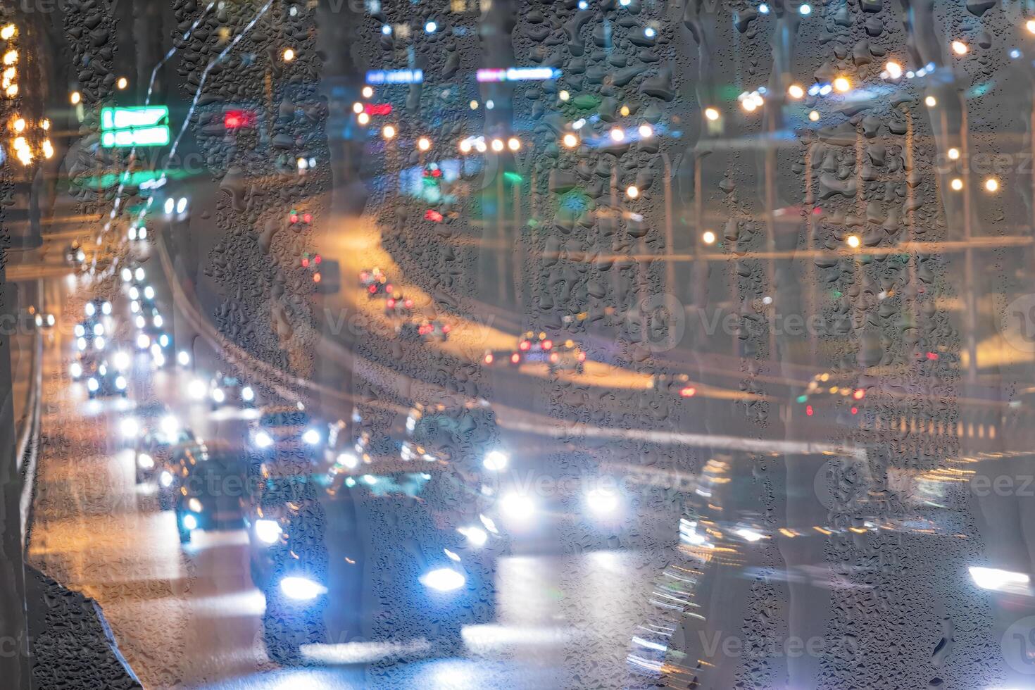 Blurry moving cars with headlights and lanterns reflecting on the wet asphalt in the night city behind rain-covered window. photo