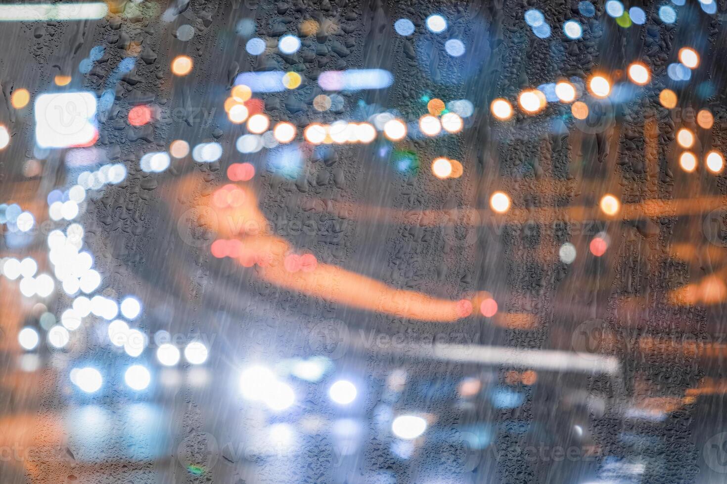 Blurry moving cars with headlights and lanterns reflecting on the wet asphalt in the night city behind rain-covered window. photo