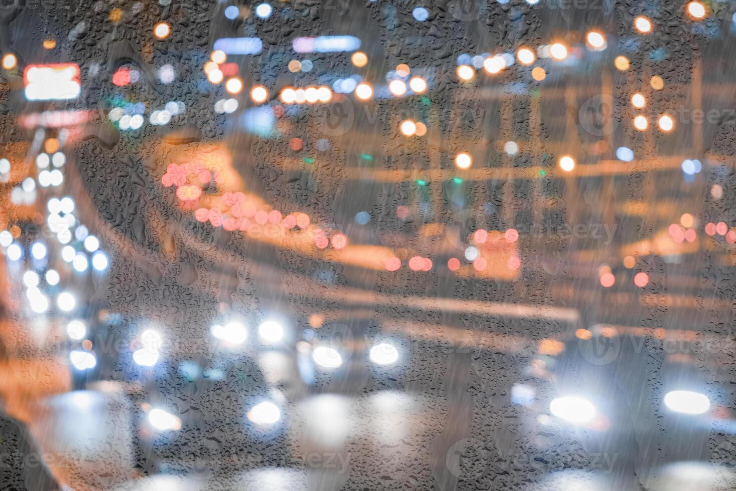 Blurry moving cars with headlights and lanterns reflecting on the wet asphalt in the night city behind rain-covered window. photo
