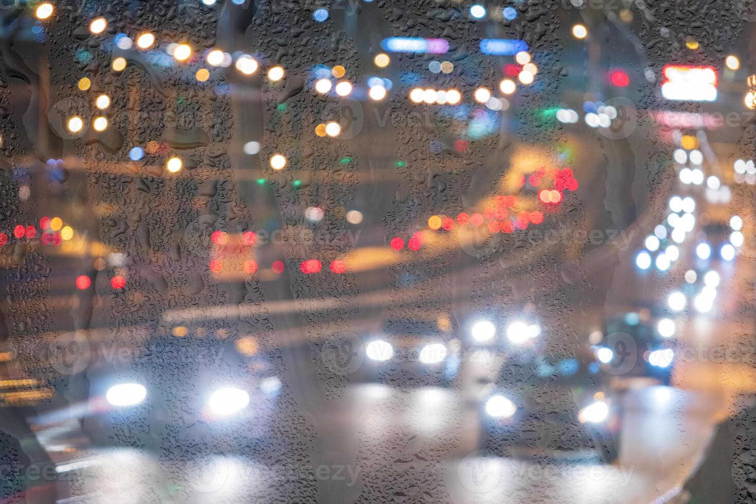 Blurry moving cars with headlights and lanterns reflecting on the wet asphalt in the night city behind rain-covered window. photo