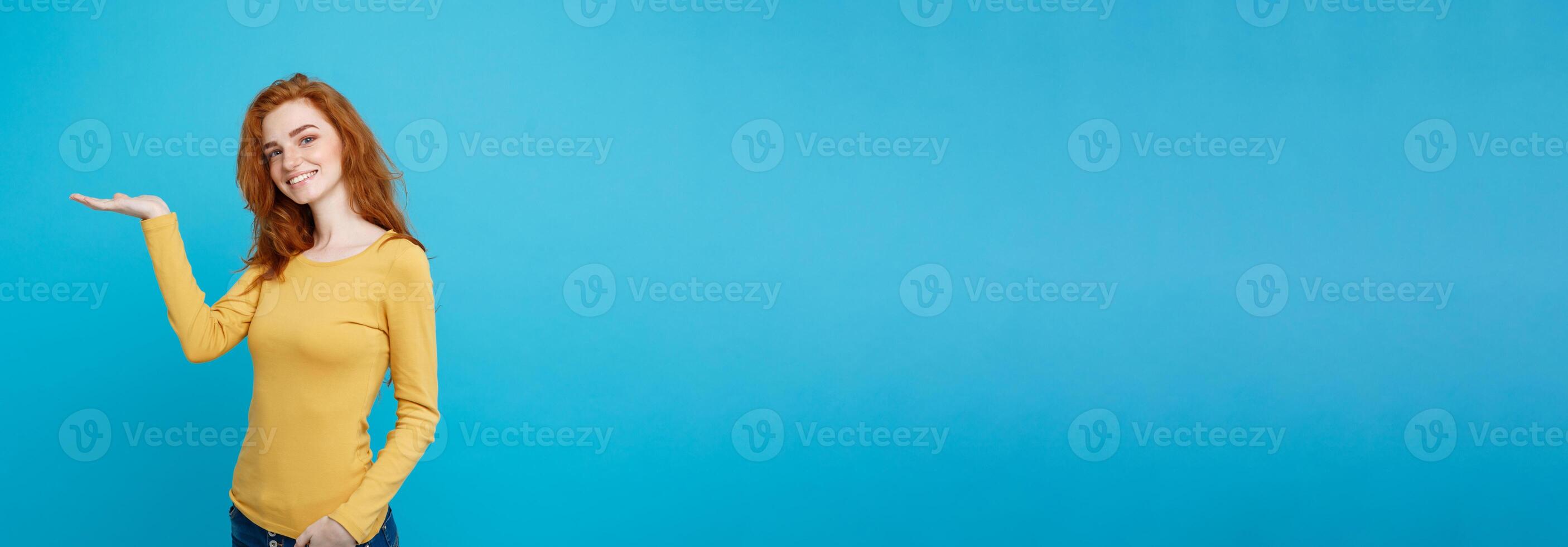 Fun and People Concept - Headshot Portrait of happy ginger red hair girl with presenting hand away and happy expression. Pastel blue background. Copy Space. photo