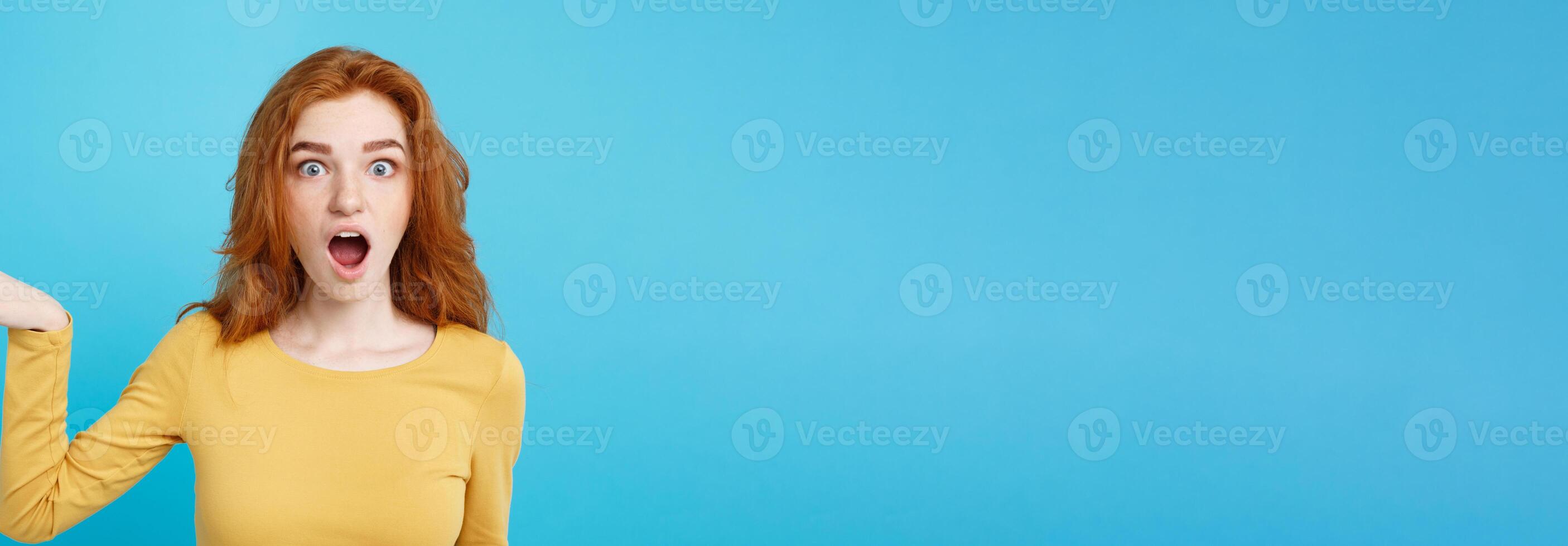 Fun and People Concept - Headshot Portrait of happy ginger red hair girl with pointing finger away and shocking expression. Pastel blue background. Copy Space. photo