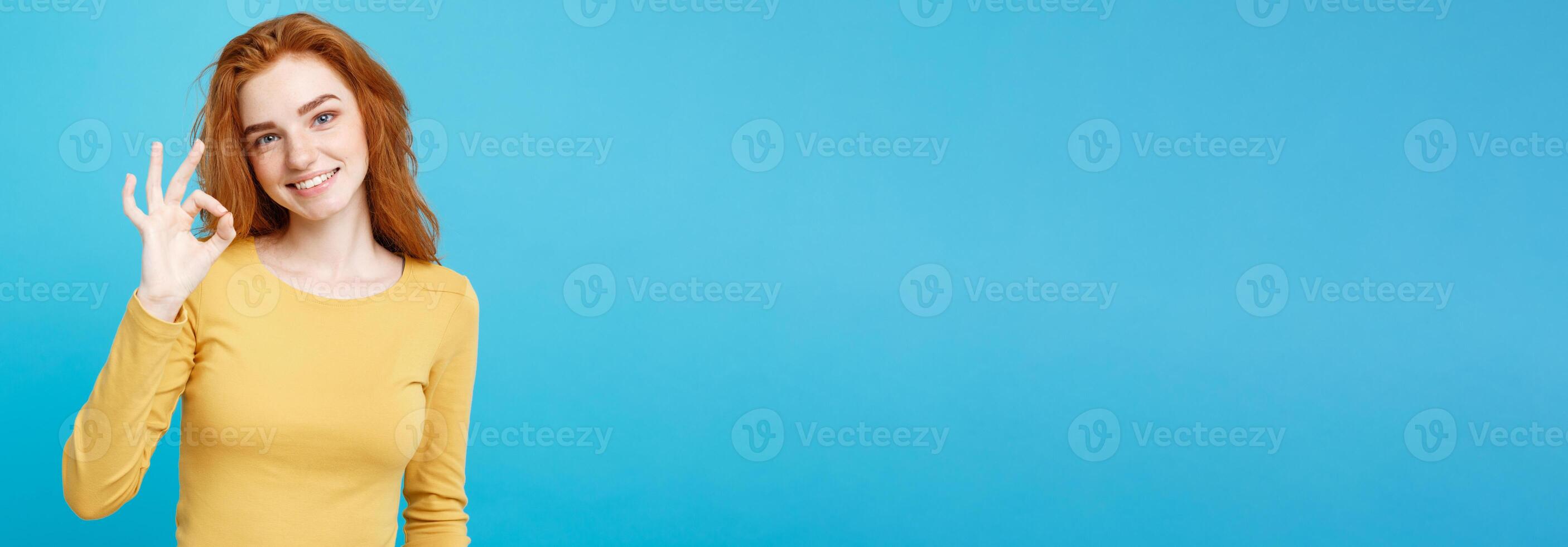 Fun and People Concept - Headshot Portrait of charming ginger red hair girl with freckles smiling and making ok sign with finger. Pastel blue background. Copy Space. photo