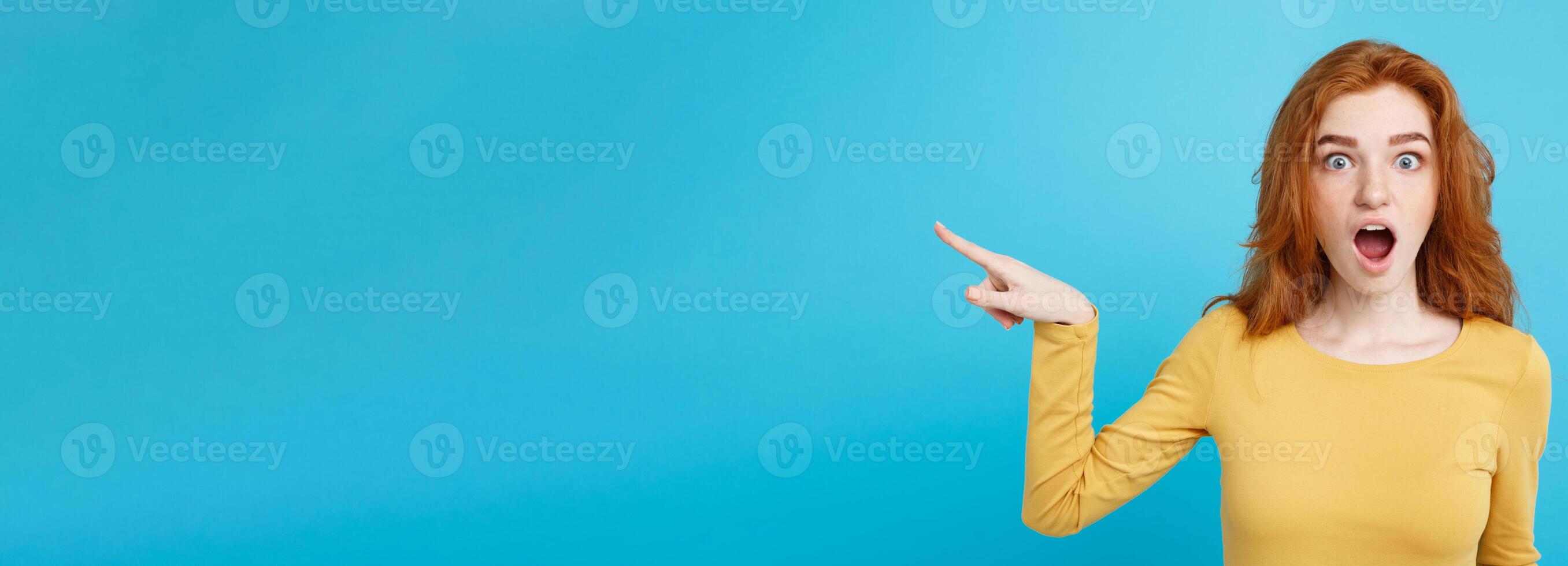 Fun and People Concept - Headshot Portrait of happy ginger red hair girl with pointing finger away and shocking expression. Pastel blue background. Copy Space. photo