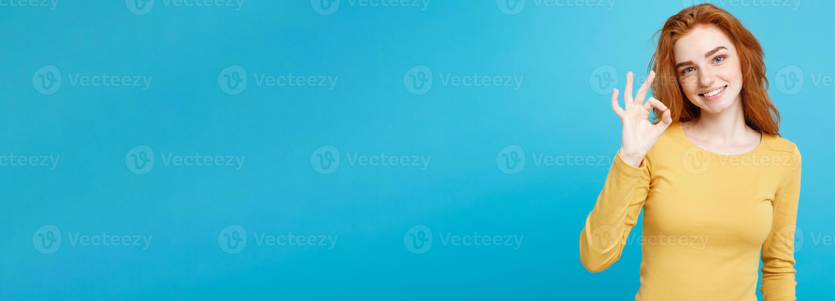 Fun and People Concept - Headshot Portrait of charming ginger red hair girl with freckles smiling and making ok sign with finger. Pastel blue background. Copy Space. photo