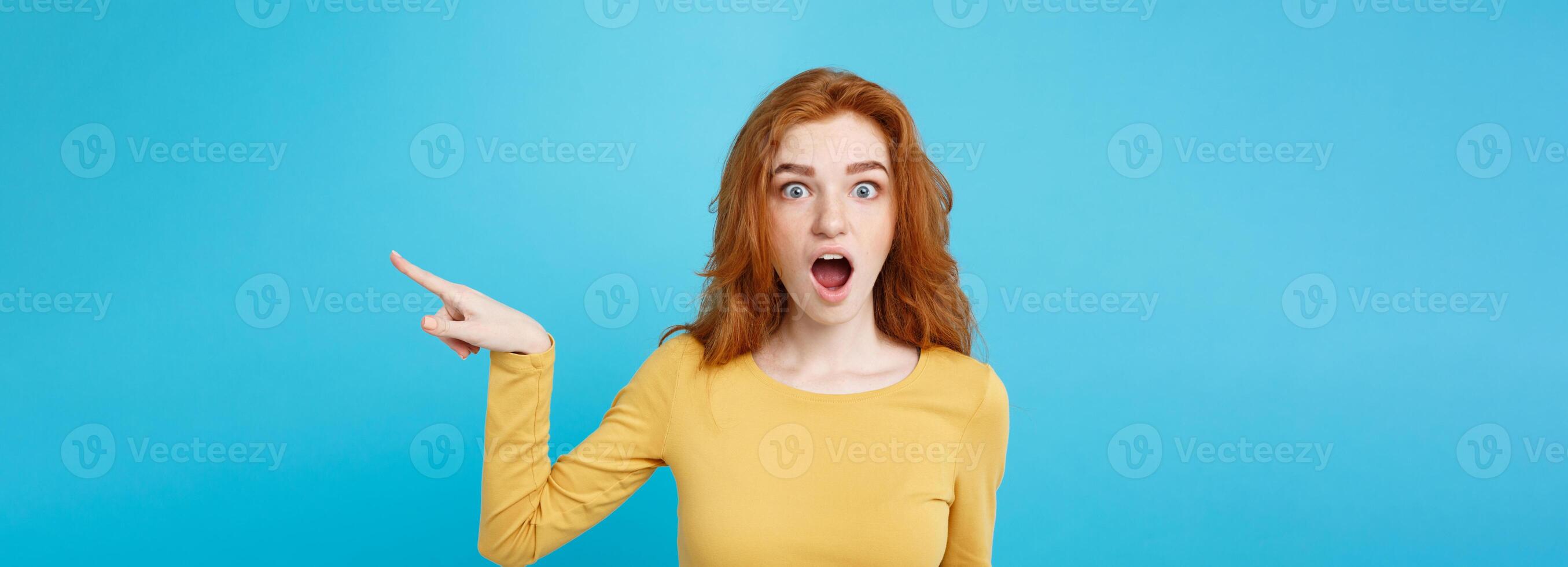 Fun and People Concept - Headshot Portrait of happy ginger red hair girl with pointing finger away and shocking expression. Pastel blue background. Copy Space. photo