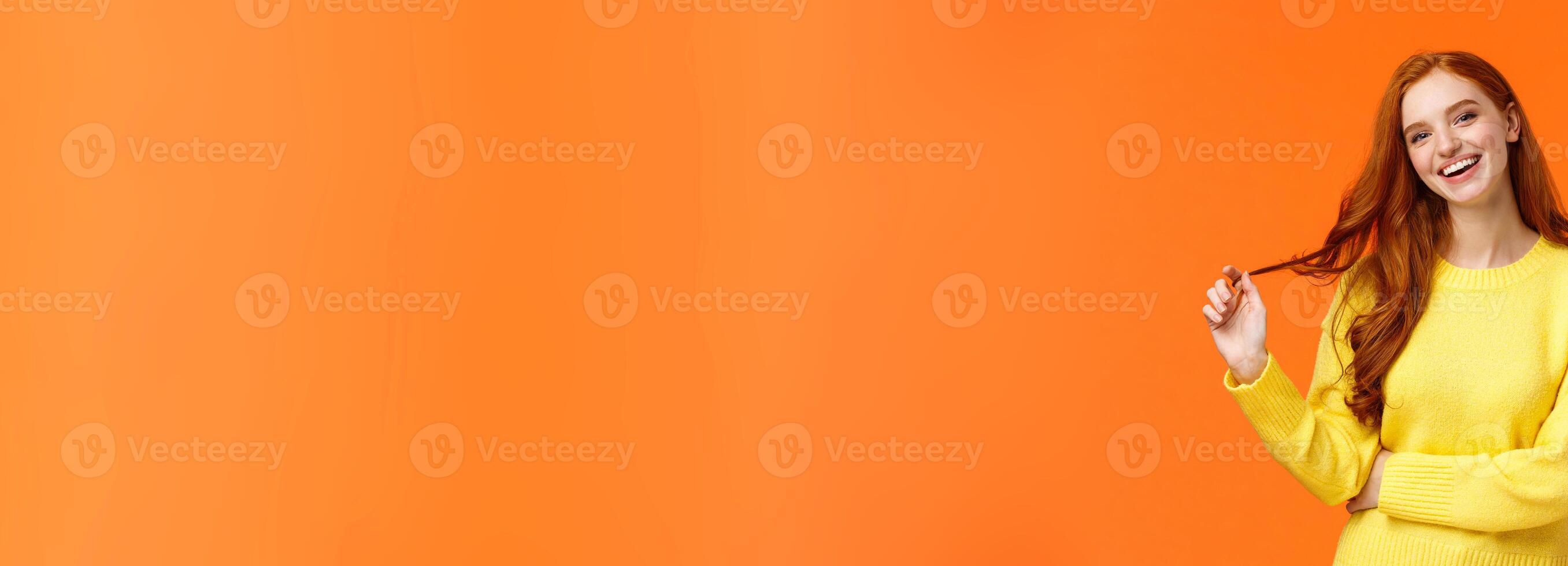 Carefree good-looking, smiling toothy redhead woman, girl with ginger hair laughing as chatting friends, rolling curl on hair, tilt head and smiling joyfully, having conversation, orange background photo