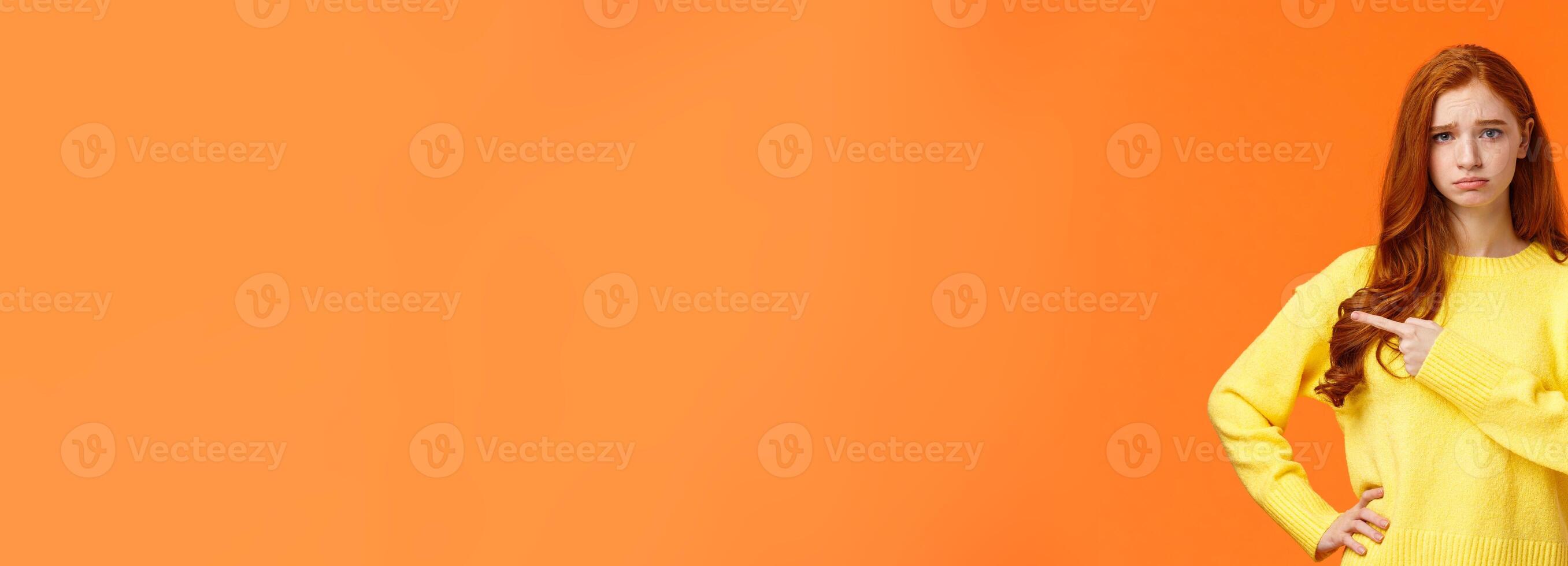 Girl looking upset and unsatisfied. Sad sulky redhead woman pointing left with envy or jealousy cant make it to party, frowning and sulking, showing what she want, standing orange background unhappy photo