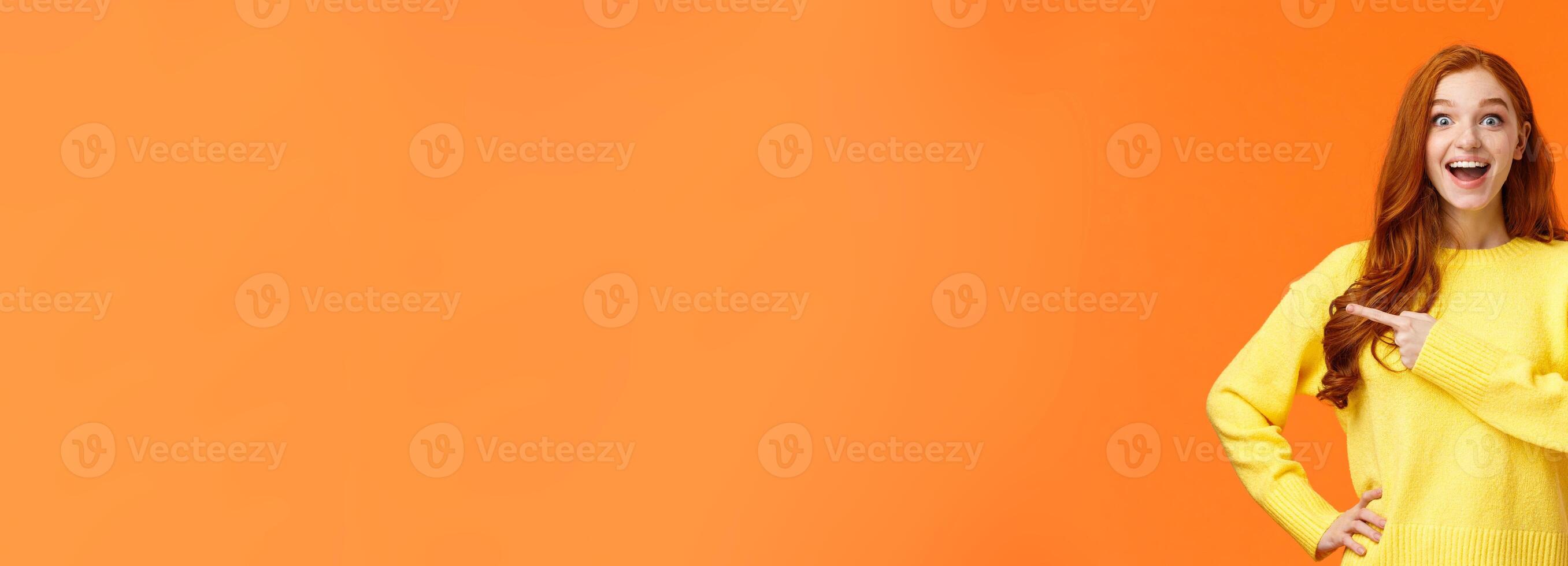 Happy, upbeat and dreamy cute redhead woman got chance travel abroad, telling about awesome company, promoting product on sale, stare fascinated camera smiling and pointing left, orange background photo