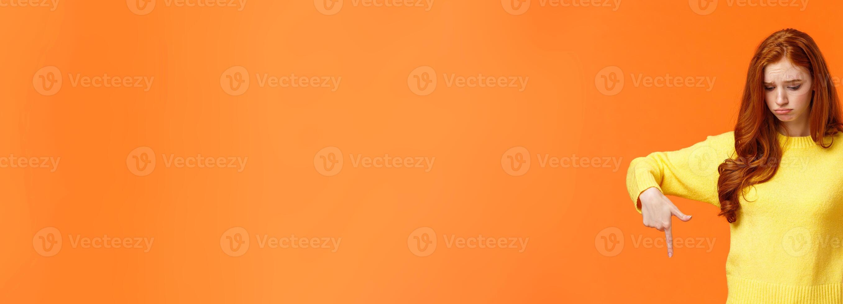 Envy, sad and sulky cute redhead woman in yellow sweater, looking and pointing down with jealousy or disappointment, feeling jealousy and regret missing chance, standing orange background photo
