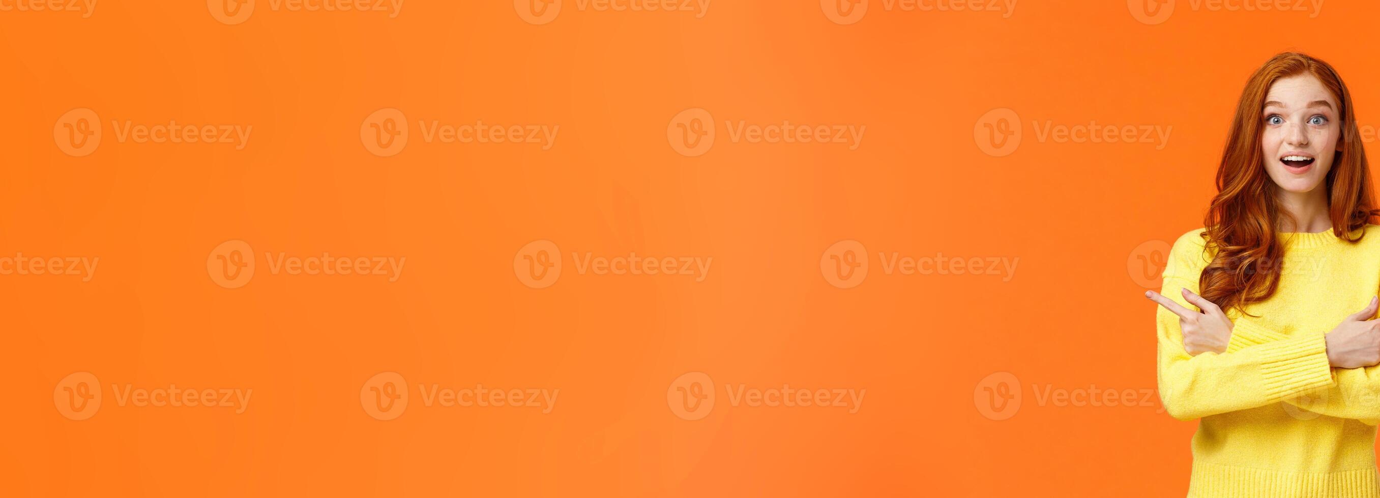 Amused dreamy and cute redhead girl asking advice in shopping mall, buying holiday presents, want valentines day gift, pointing sideways left and right, smiling upbeat, orange background photo
