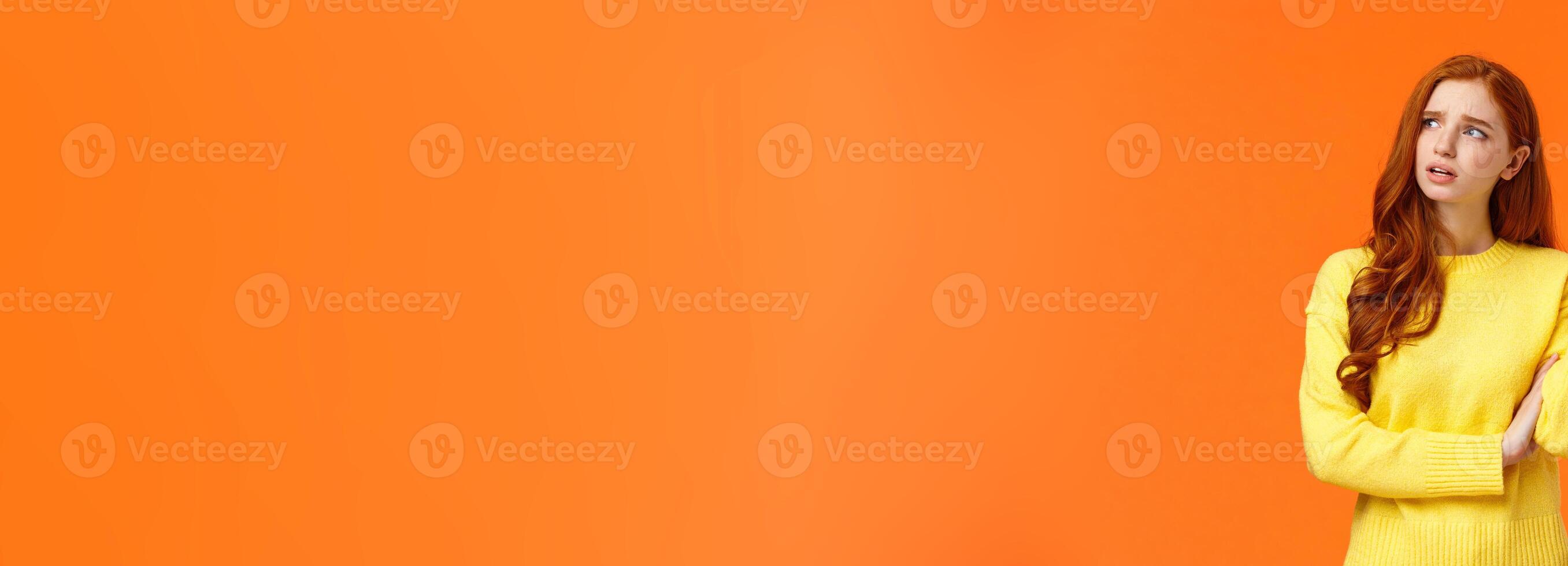 Troubled and unsure, indecisive cute redhead girl trying solve problem, searching resolve, looking perplexed upper left corner pondering, raise hand uncertain, confused, shrugging, orange background photo