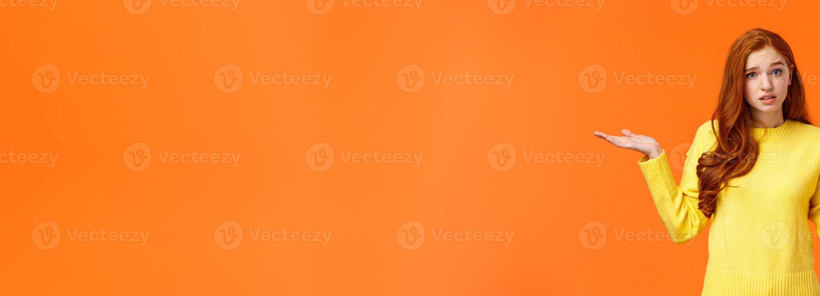 Perplexed cute redhead girl, shrugging with raised arms and looking confused, dont know answer, cant help, feeling confused or indecisive, cannot understand, standing orange background photo