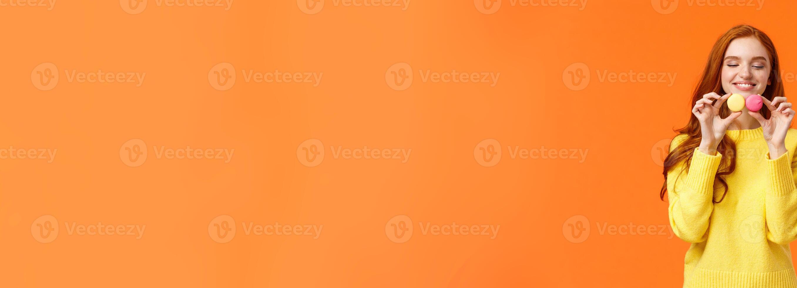 Redhead silly and happy gorgeous girl buy tasty macarons, holding food and looking with desire and affection, like sweets, want eat them, standing orange background satisfied and joyful photo