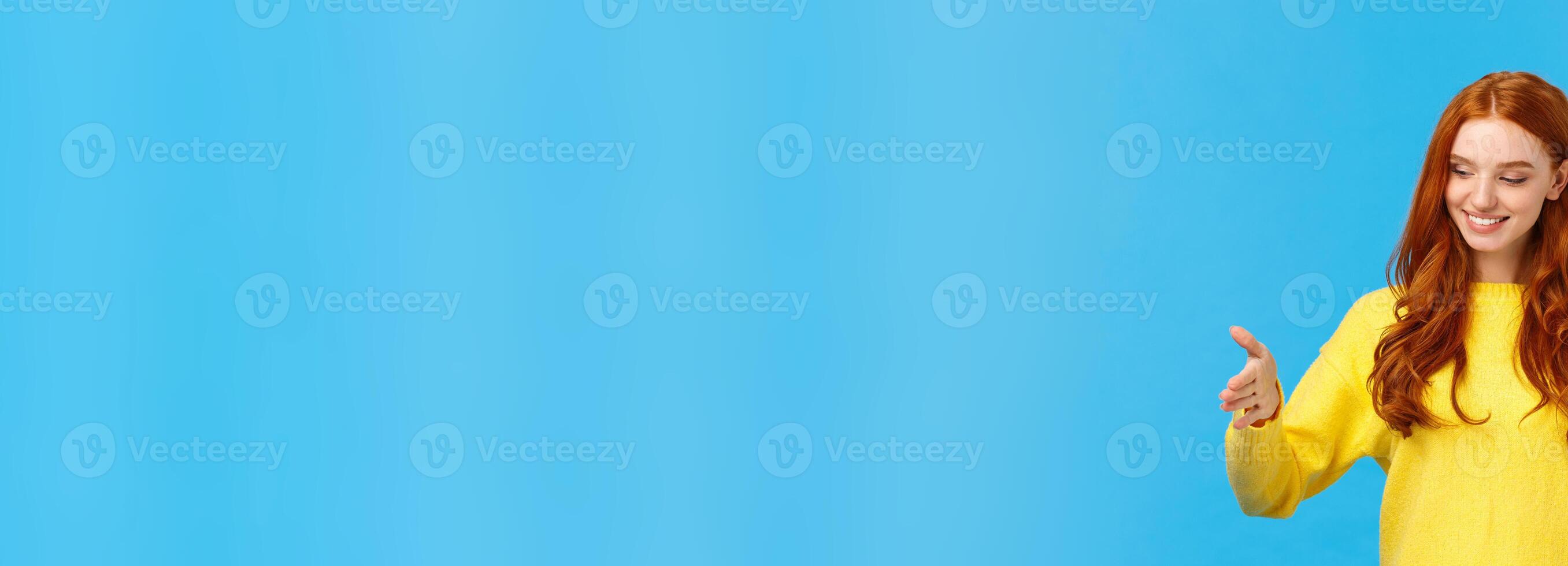Happy good-looking young woman with red curly hair, freckles, shaping long object as if holding box over chest, looking satisfied, pleased with size, explain how big was gift, blue background photo