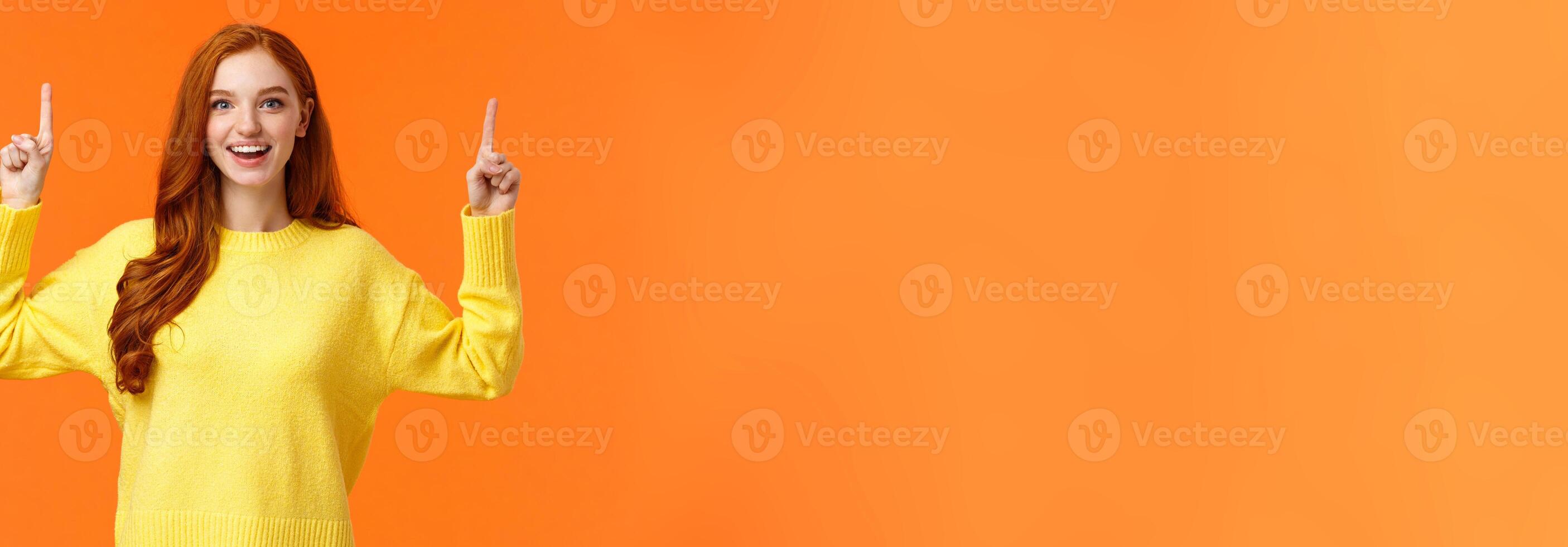 Excited, tender good-looking redhead girl with freckles, wear yellow sweater, pointing up and smiling joyfully, telling about promotion, advertising top product, orange background photo