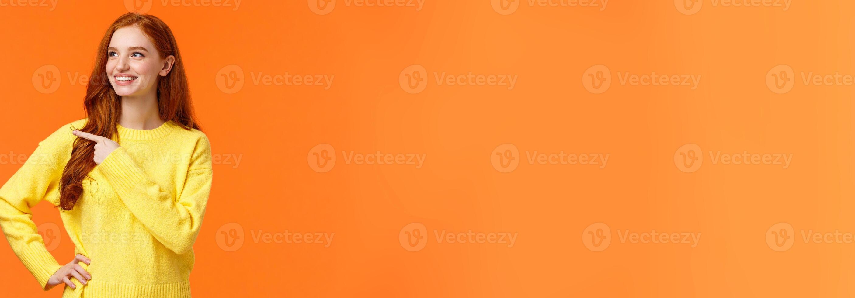 Dreamy, energized good-looking redhead female in yellow sweater, found excellent holiday gift, consumer pointing left, looking satisfied, smiling as picking product, make decision, orange background photo