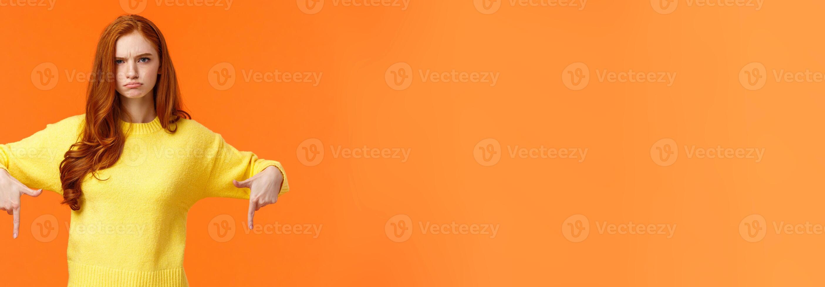 Mad unsatisfied cute redhead girlfriend, ginger girl in yellow sweater, frowning disappointed and angry, pointing down sulking, feeling offended with disapproval, standing orange background photo