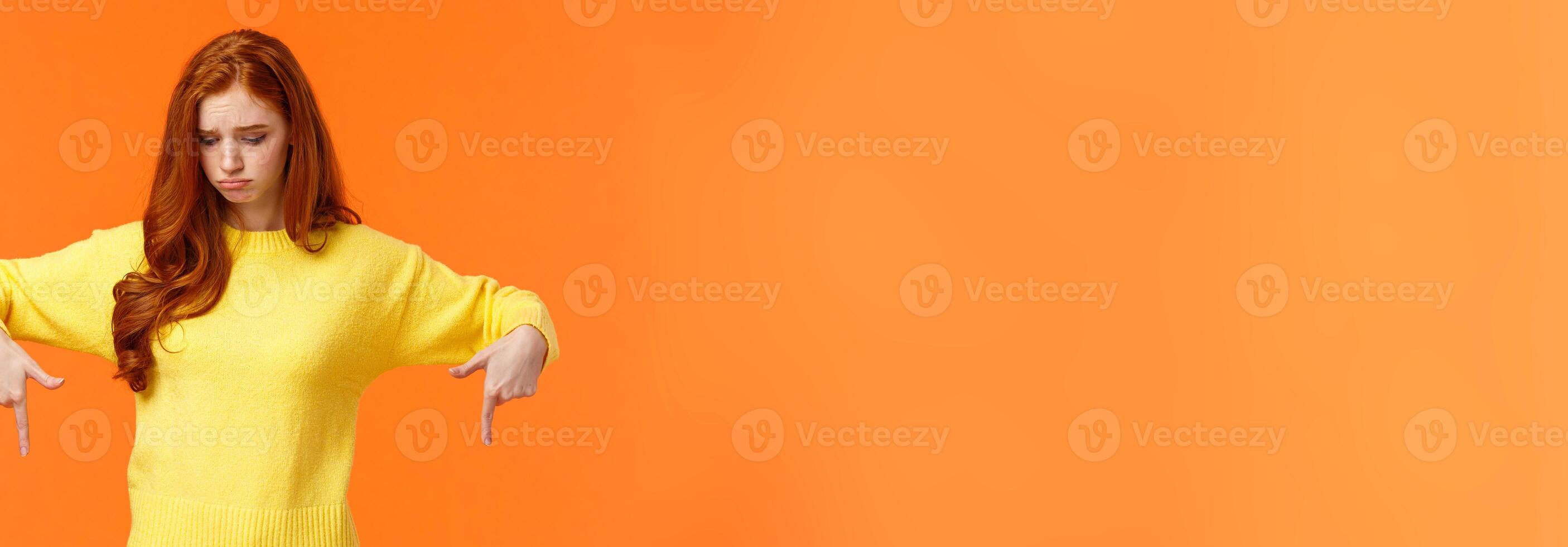 Envy, sad and sulky cute redhead woman in yellow sweater, looking and pointing down with jealousy or disappointment, feeling jealousy and regret missing chance, standing orange background photo