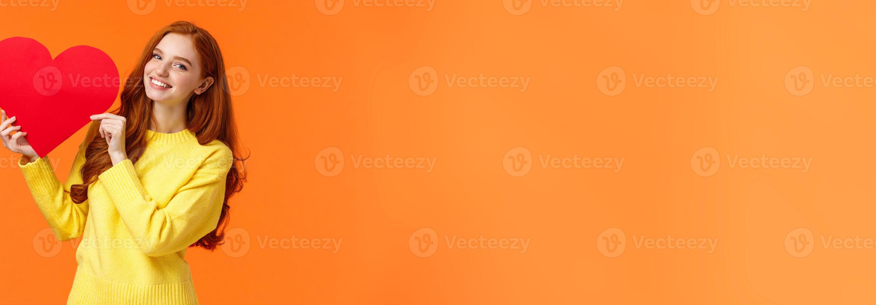 Valentines day perfect time to confess. Cute romantic and tender redhead woman in yellow sweater holding large heart sign and smiling, express affection and romance, standing orange background photo