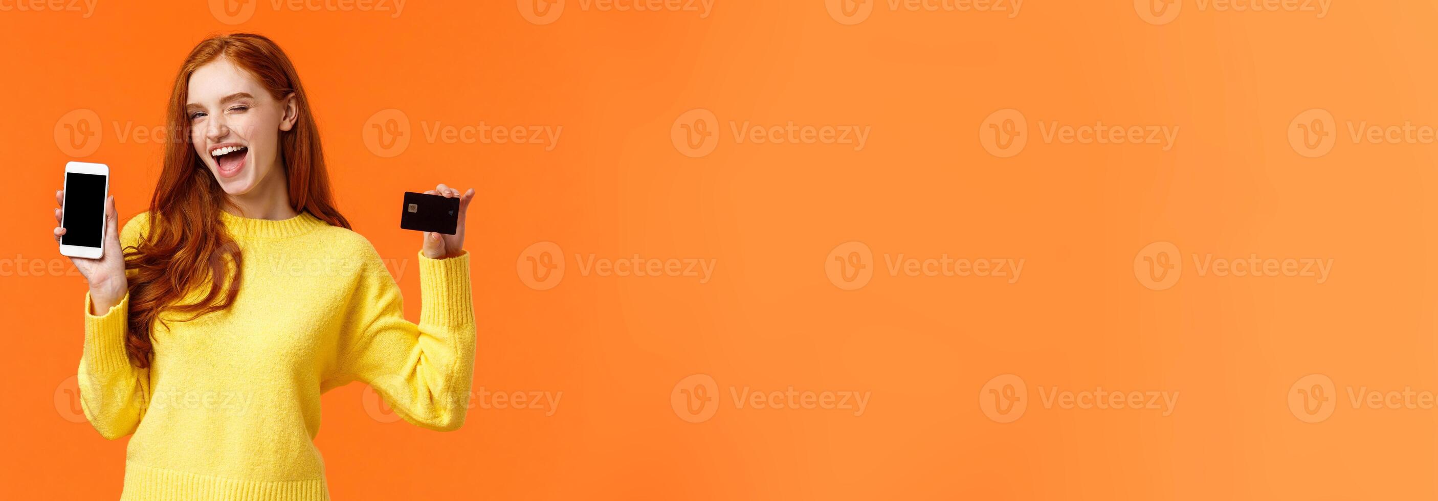 Shopping, advertising and mobile addication concept. Attractive cheeky redhead girl showing smartphone and credit card, wink and smile camera, promote online banking, deposit or payment method photo