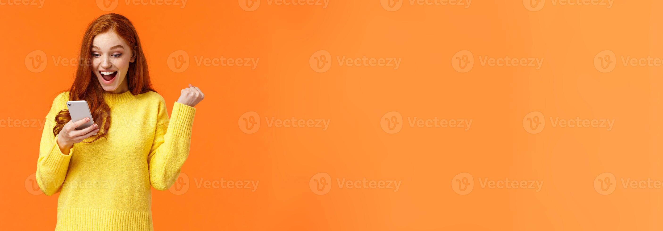 Girl received thousand likes under new picture. Cheerful and triumphing pretty redhead female fist pump say yes, cheering or celebrating good news, looking smartphone screen, orange background photo