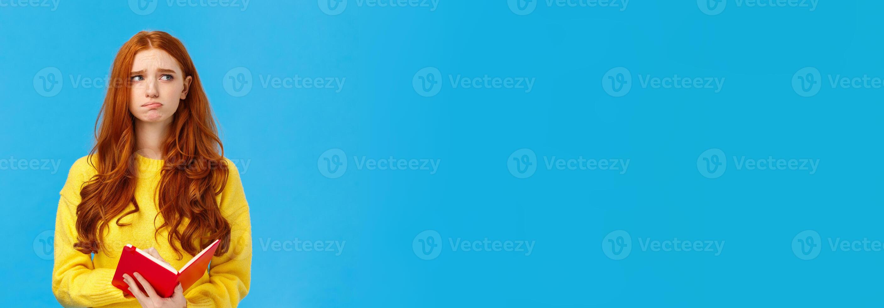 Troubled and pensive uneasy pretty redhead female, pouting and frowning looking away thoughtful, feeling sad and distressed, reading something bad in notebook, standing blue background photo
