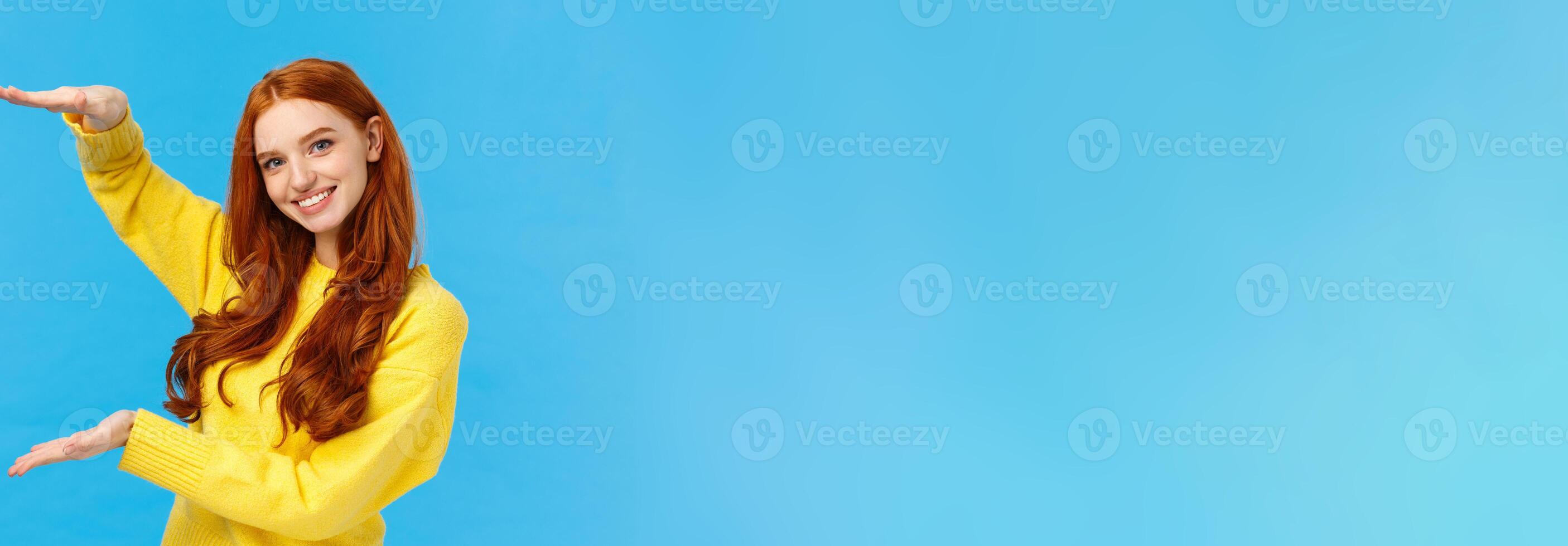 Cute friendly-looking caucasian redhead woman shaping large object with hands, presenting gift, product advertising over blue background, smiling pleasantly, bragging big birthday present photo
