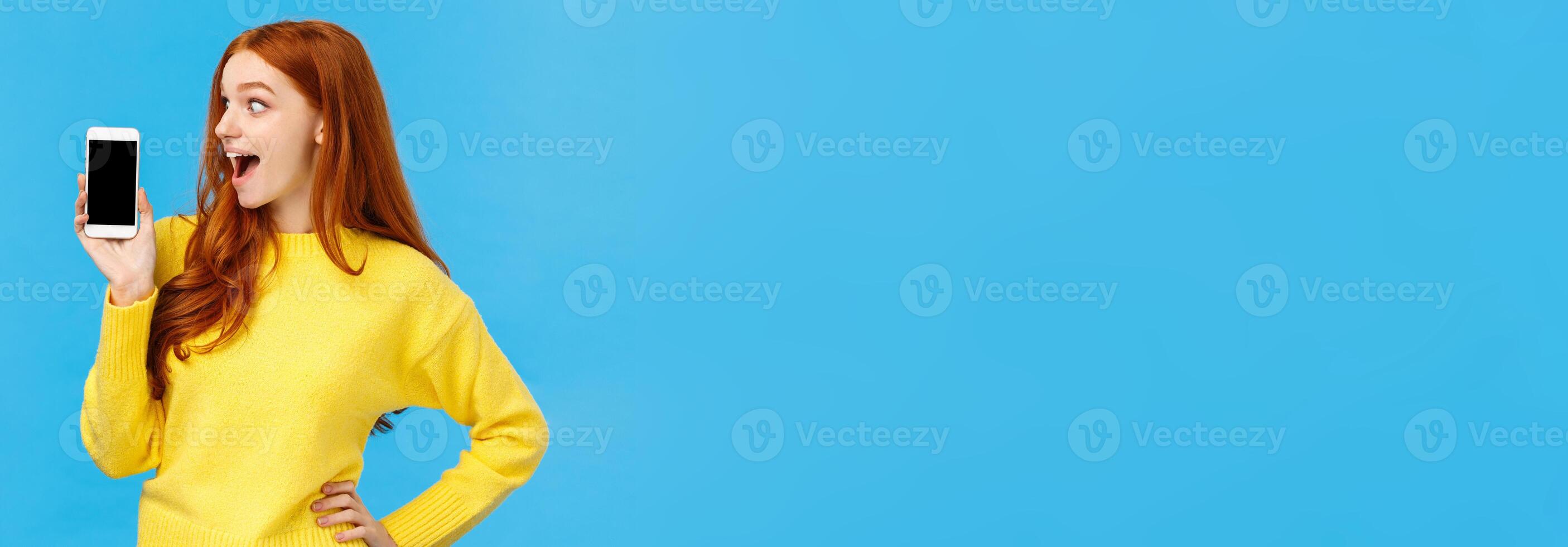 Wow look at this app. Amused and wondered, excited redhead girl looking at smartphone display, showing mobile screen, shopping site or application, standing blue background photo
