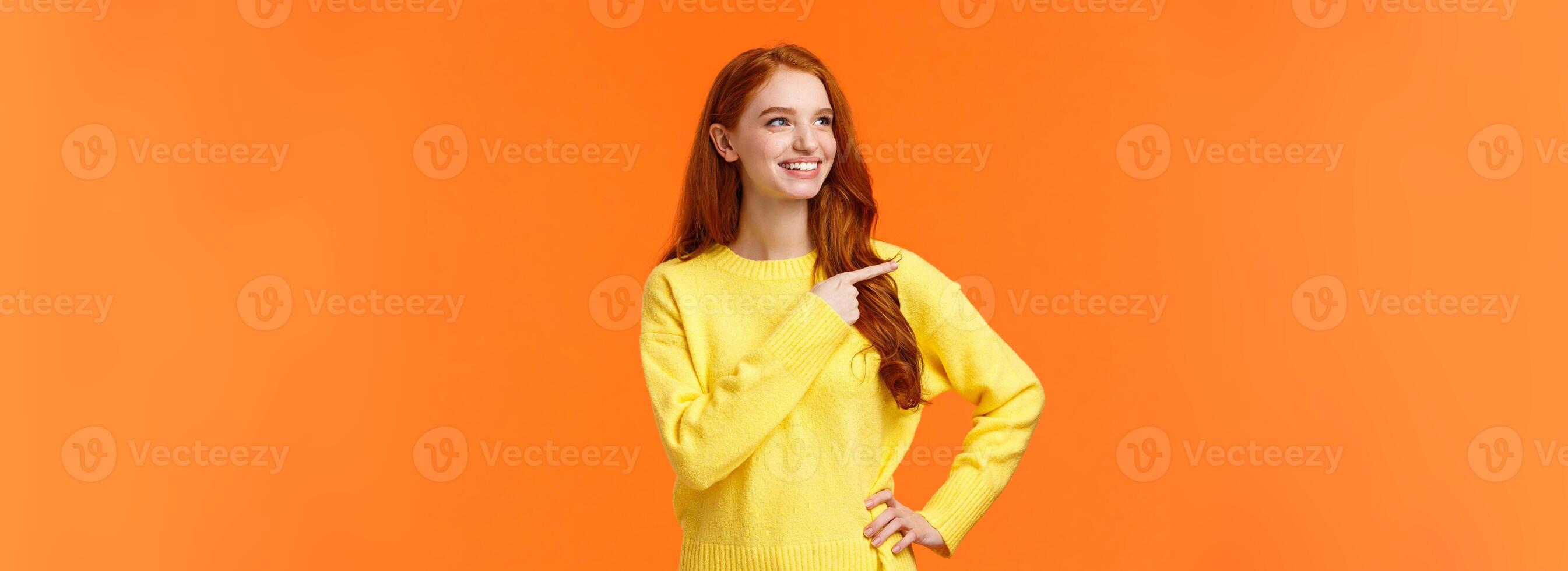 Dreamy, energized good-looking redhead female in yellow sweater, found excellent holiday gift, consumer pointing left, looking satisfied, smiling as picking product, make decision, orange background photo