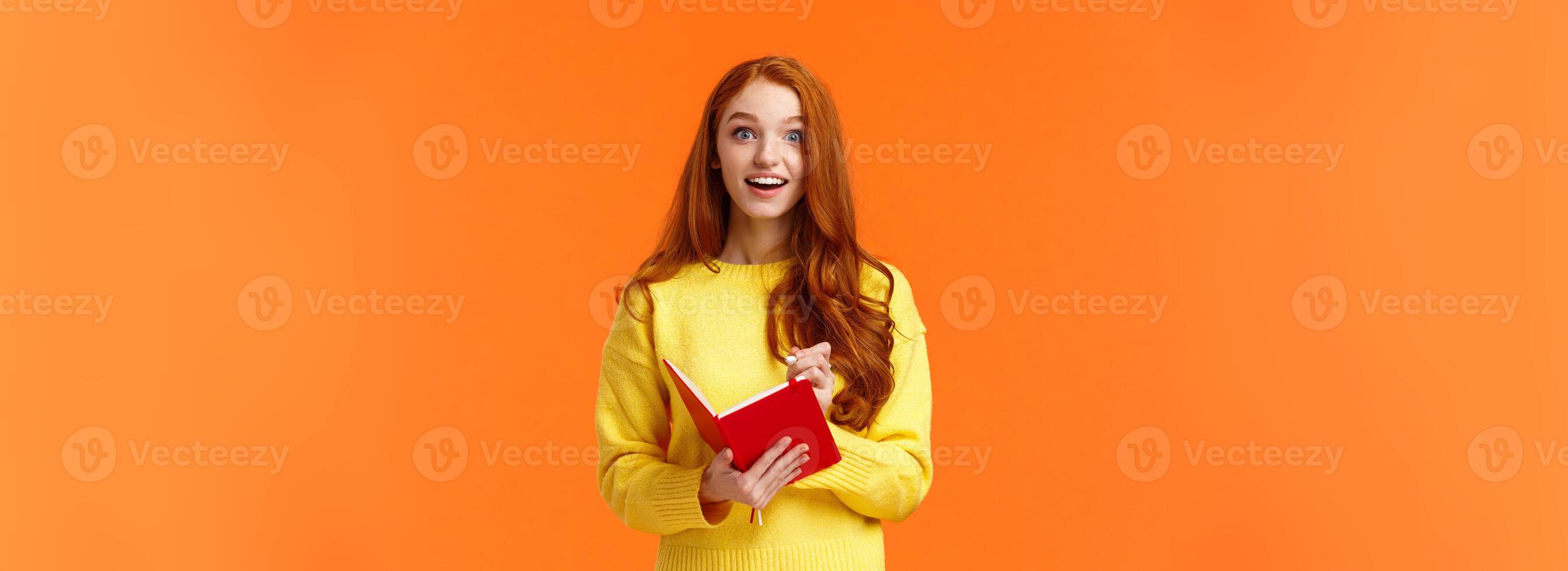 Inspired and upbeat, dreamy fascinated redhead girl writing down interesting lecture, smiling and gazing with admiration or enthusiasm, fill-in notebook schedule, standing orange background photo