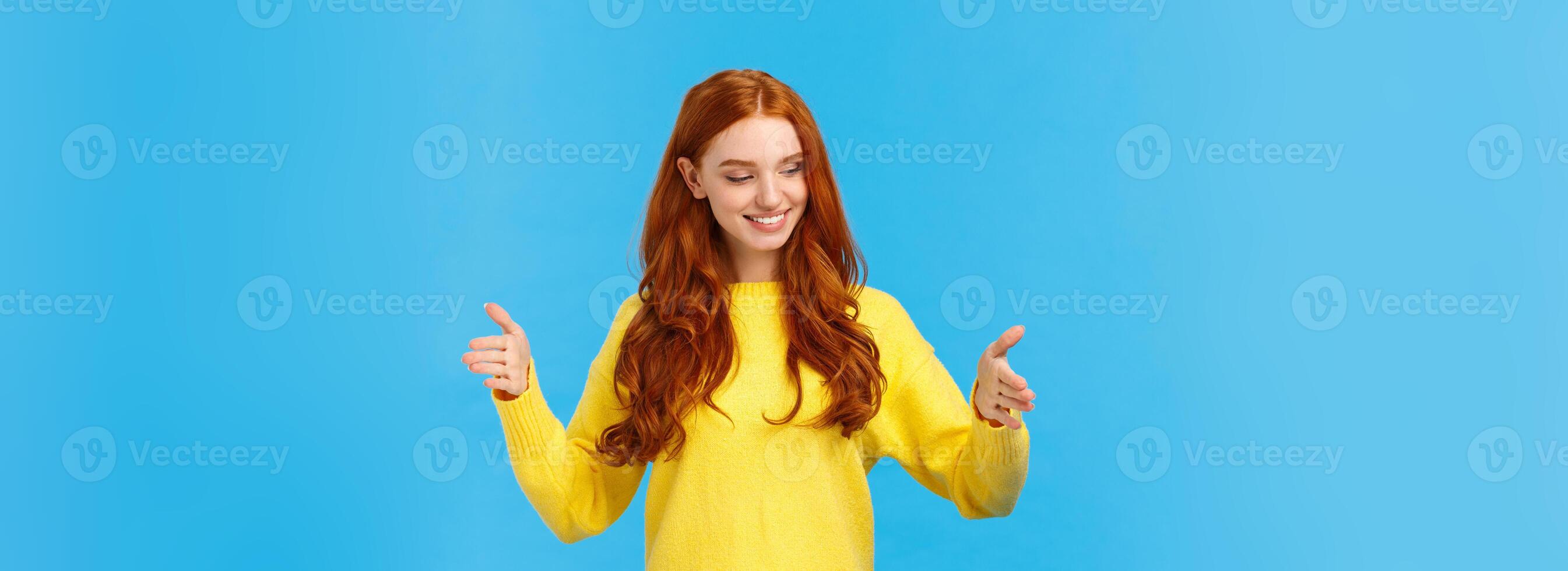 Happy good-looking young woman with red curly hair, freckles, shaping long object as if holding box over chest, looking satisfied, pleased with size, explain how big was gift, blue background photo