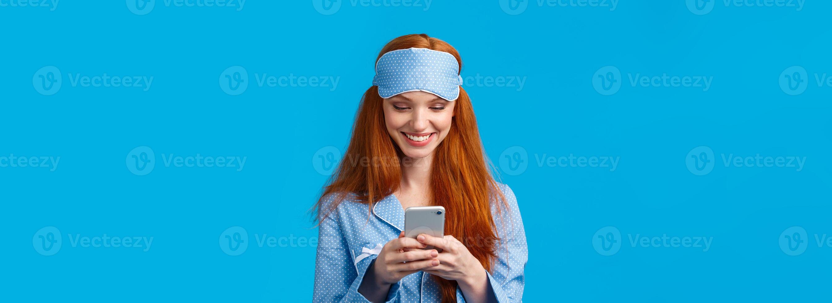 Mobile addication, technology and beauty concept. Cheerful girl waking up and grab phone, checking messages, scrolling news feed on smartphone wearing nightwear, sleep mask, smiling photo