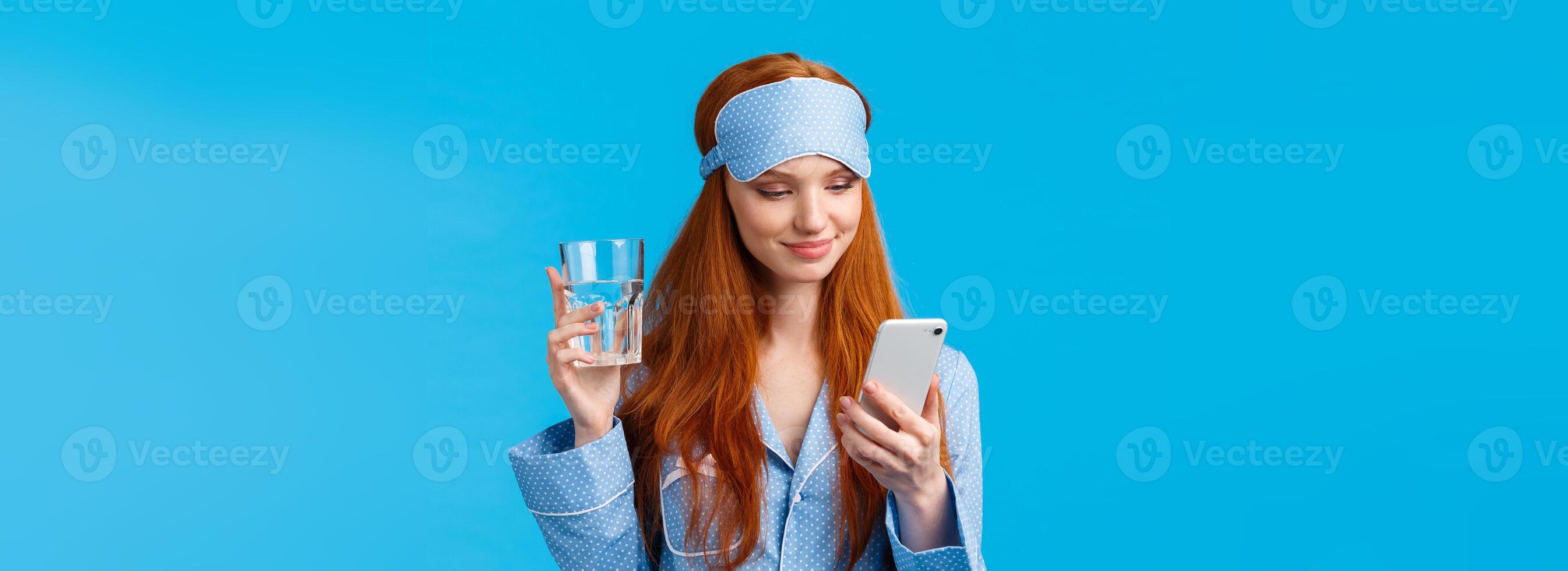 Waist-up portrait joyful redhead european glamour girl in sleep mask and pyjama, holding glass water and checking drinking schedule app, smiling as chatting via smartphone, blue background photo