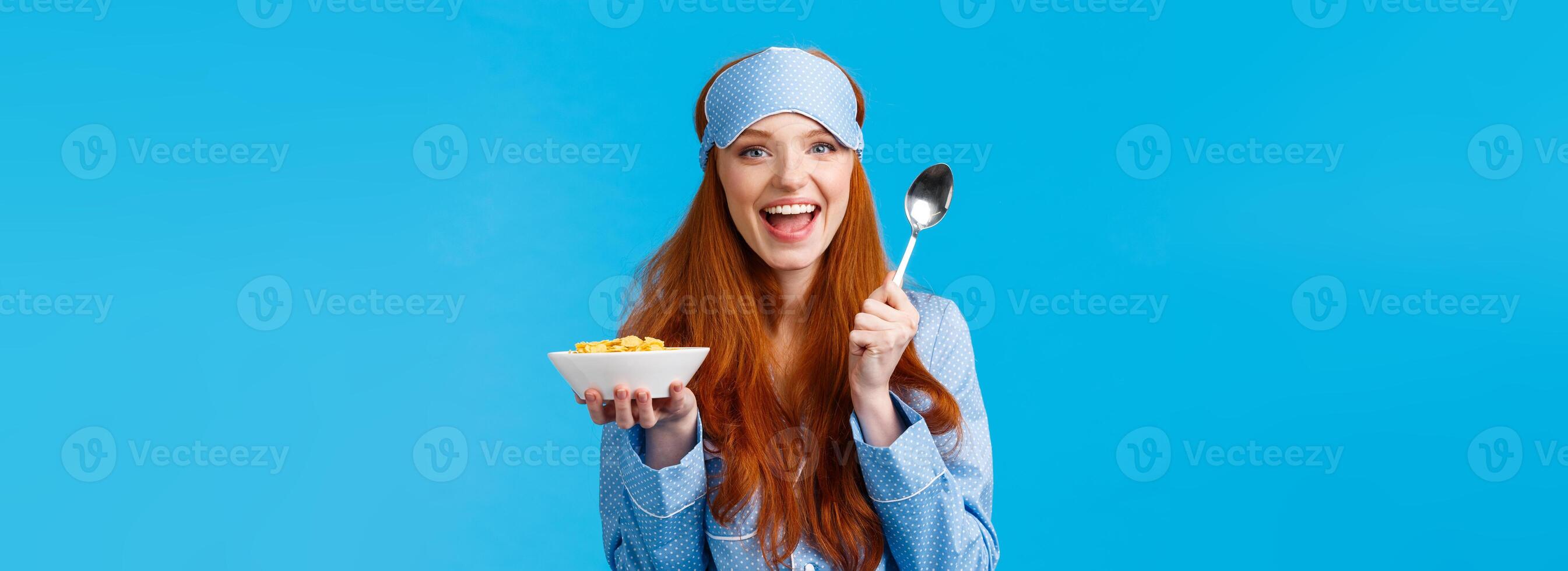How about morning breakfast. Cheerful good-looking redhead female with excited expression, smiling eating cereals and holding spoon, standing blue background in nightwear, sleep mask photo