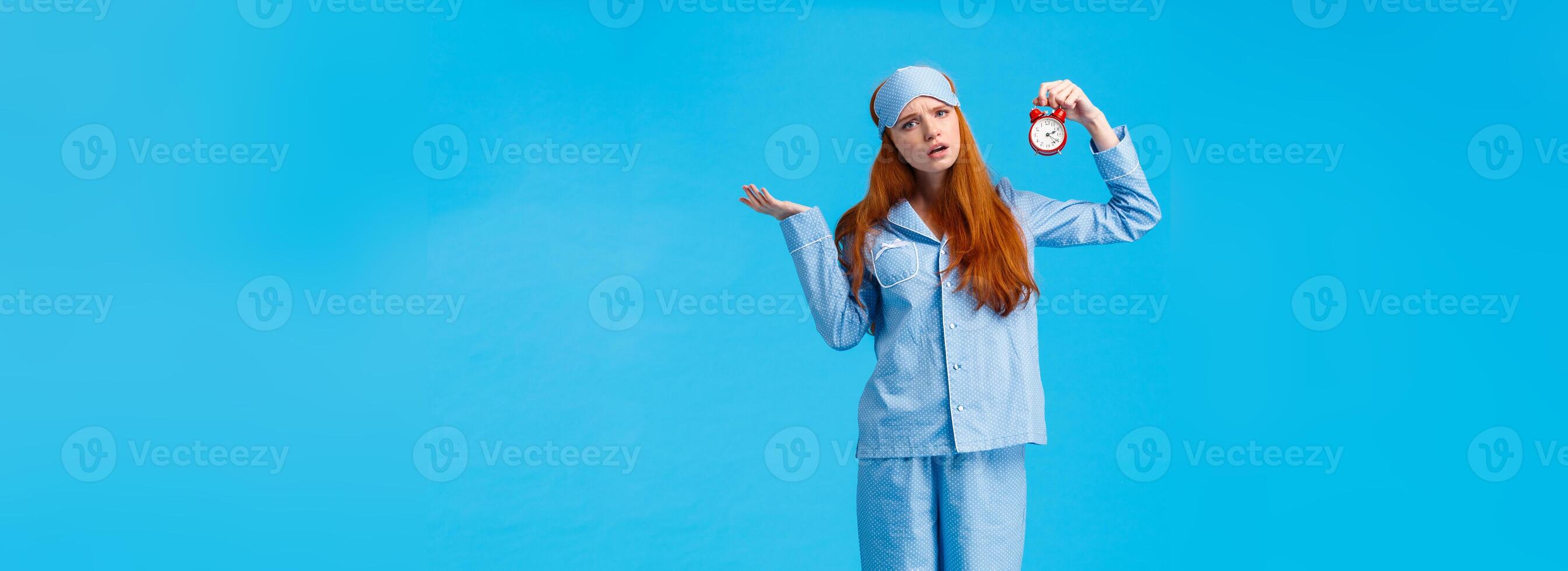 Why didnt you set alarm. Confused and unsure upset attractive redhead woman in pyjama and sleep mask, shrugging perplexed staring questioned camera, holding clokc, blue background photo