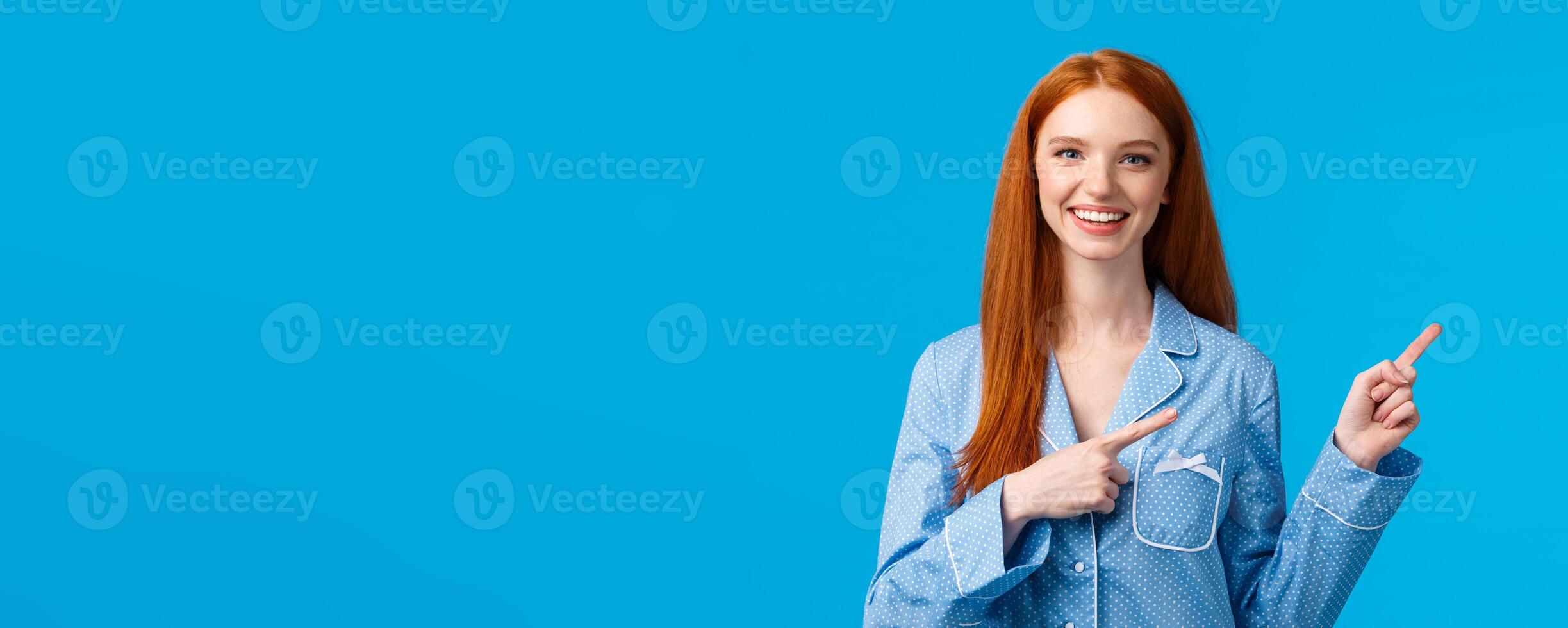Friendly attractive redhead female student showing person way in dormitory college, pointing right and smiling wearing nightwear, going sleep, showing her daily skincare routine, blue background photo