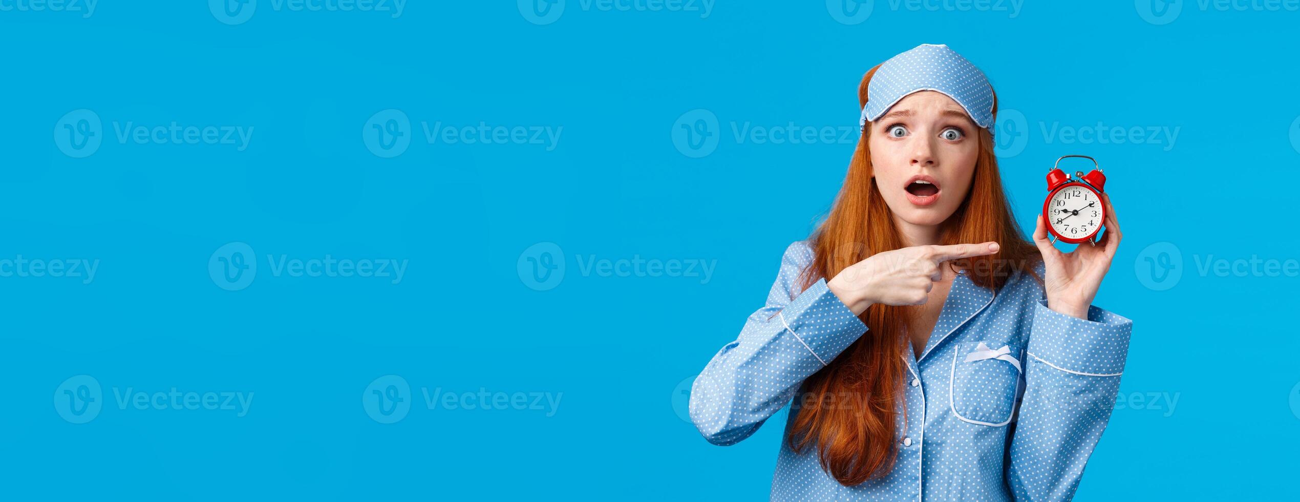Shocked and concerned, worried cute redhead girl pointing at alarm clock with frustrated, nervous expression, being late, dont know how set up right time, standing blue background in nightwear photo