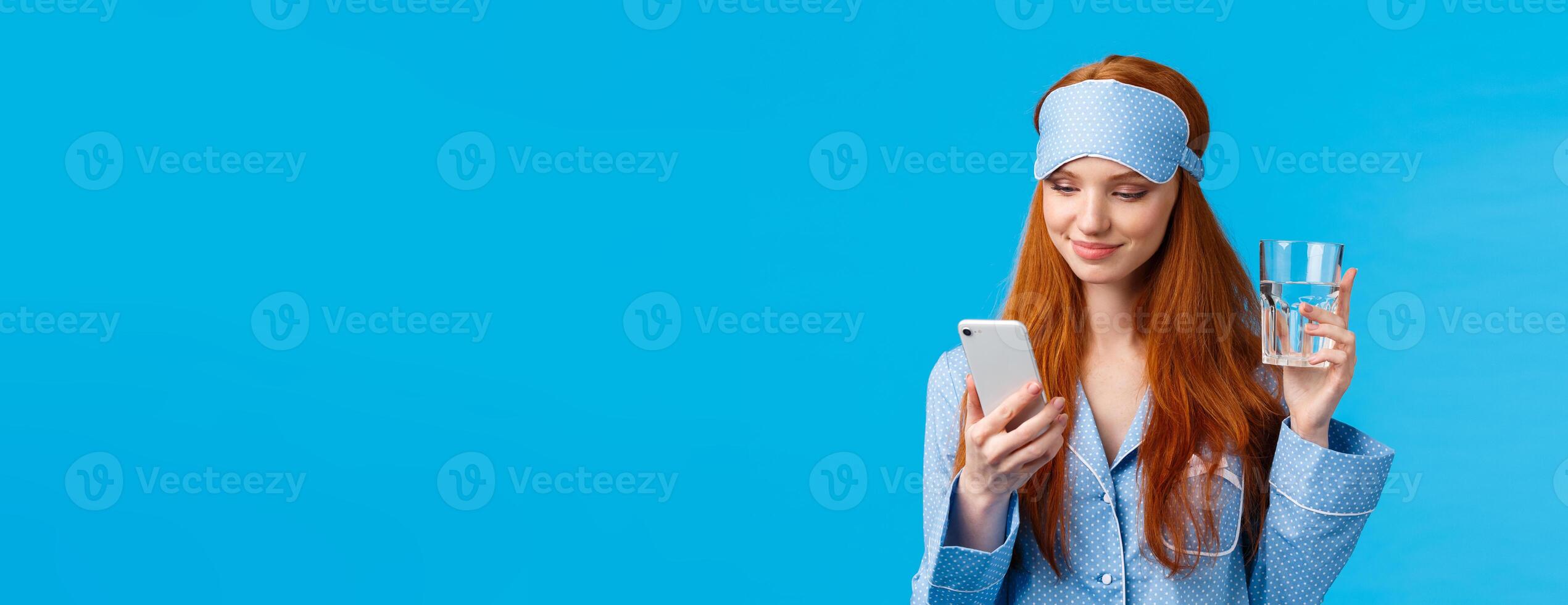 Waist-up portrait joyful redhead european glamour girl in sleep mask and pyjama, holding glass water and checking drinking schedule app, smiling as chatting via smartphone, blue background photo