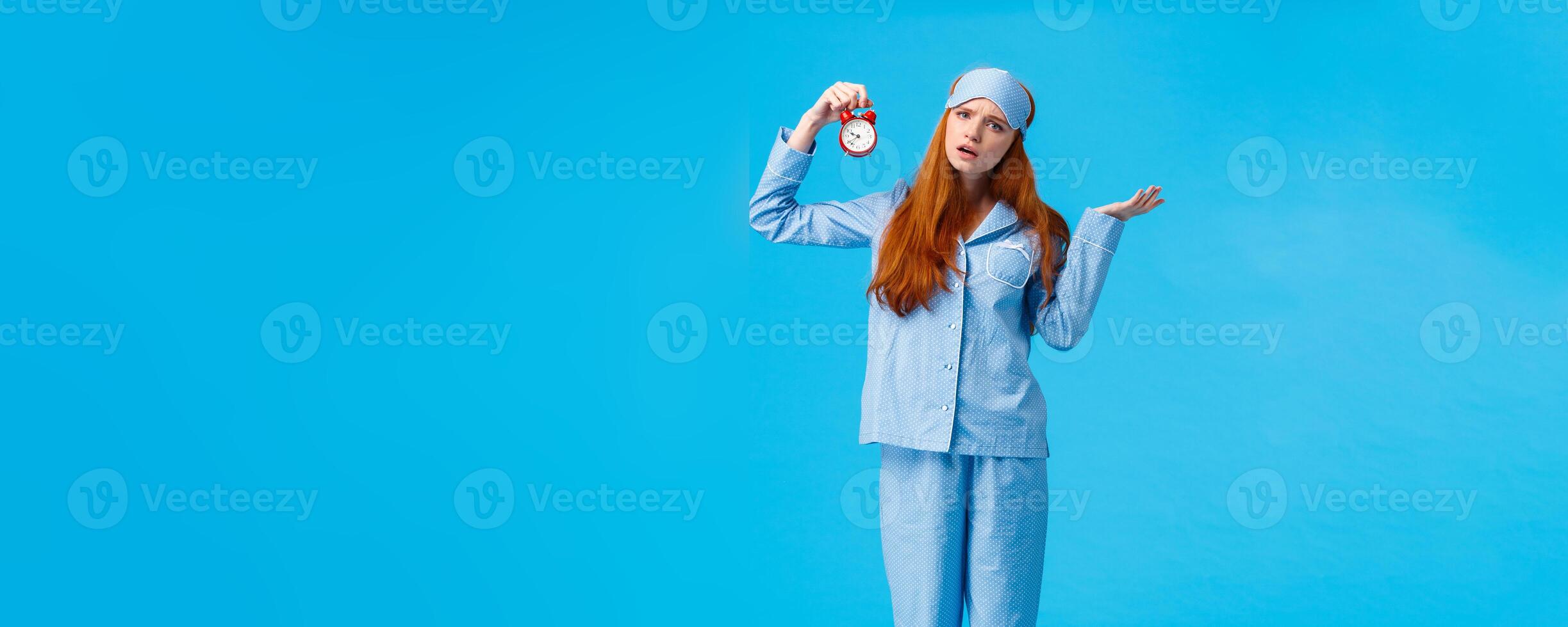 Why didnt you set alarm. Confused and unsure upset attractive redhead woman in pyjama and sleep mask, shrugging perplexed staring questioned camera, holding clokc, blue background photo
