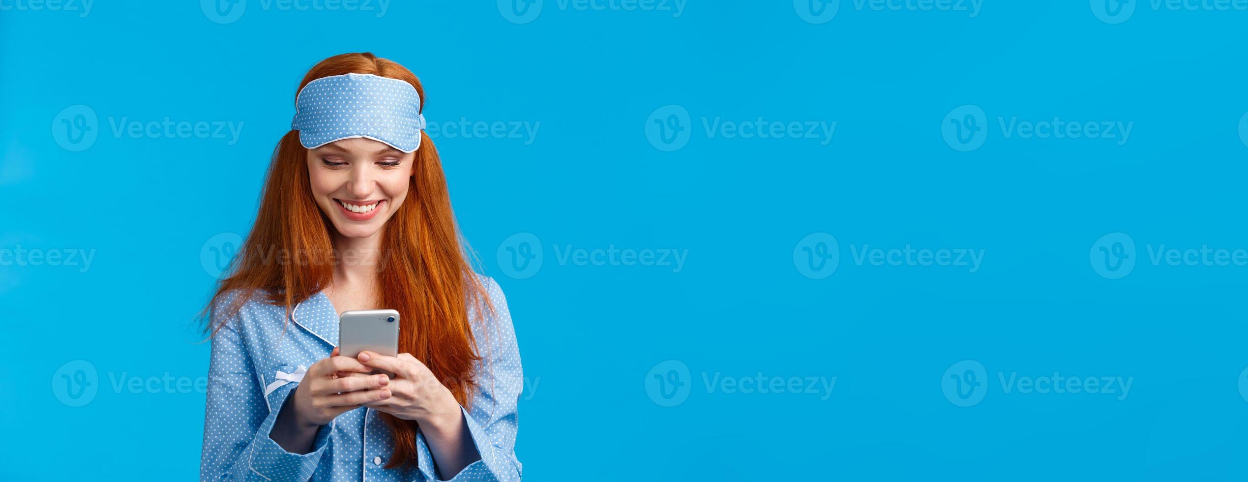 Mobile addication, technology and beauty concept. Cheerful girl waking up and grab phone, checking messages, scrolling news feed on smartphone wearing nightwear, sleep mask, smiling photo