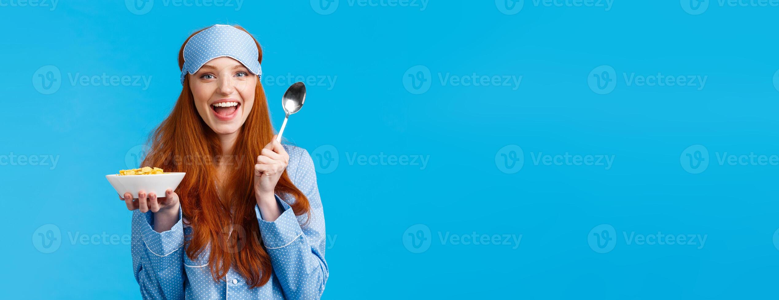 How about morning breakfast. Cheerful good-looking redhead female with excited expression, smiling eating cereals and holding spoon, standing blue background in nightwear, sleep mask photo