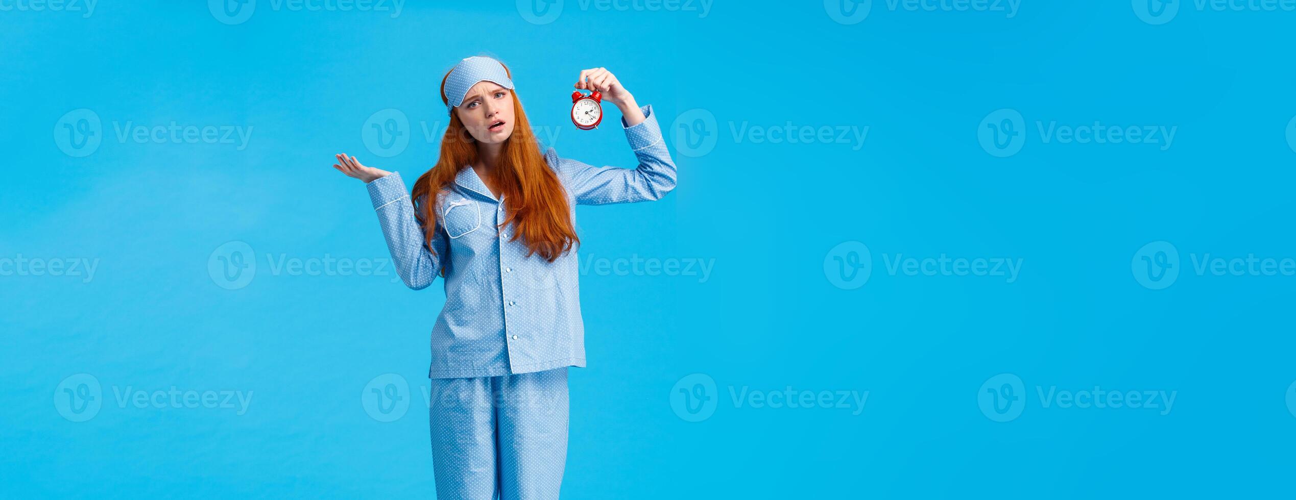 Why didnt you set alarm. Confused and unsure upset attractive redhead woman in pyjama and sleep mask, shrugging perplexed staring questioned camera, holding clokc, blue background photo