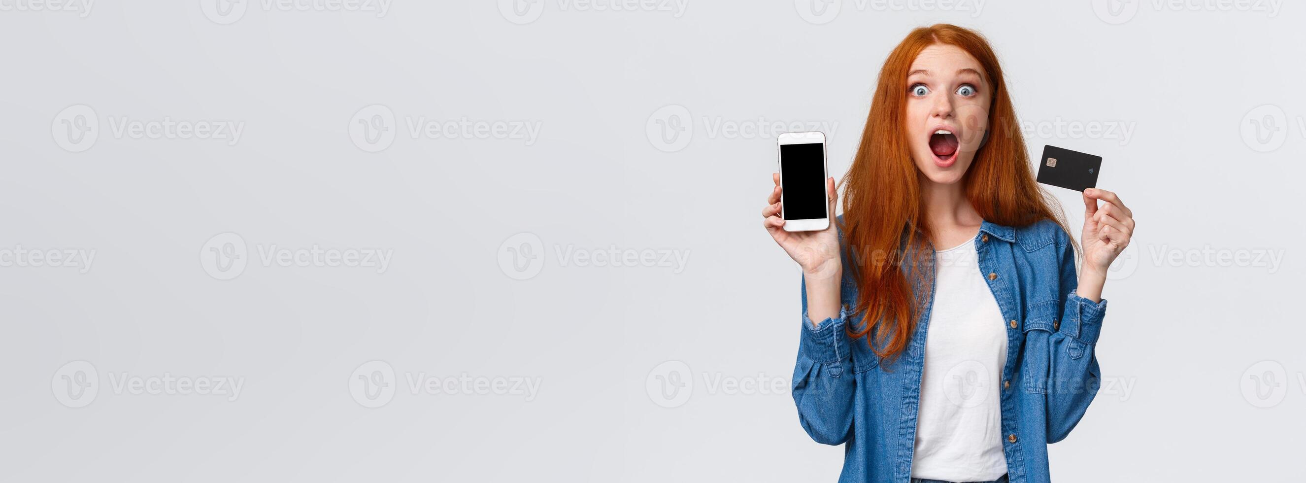 Waist-up portrait excited, astonished and thrilled redhead female student holding credit card, showing smartphone display, application, drop jaw impressed, looking camera astounded, white background photo