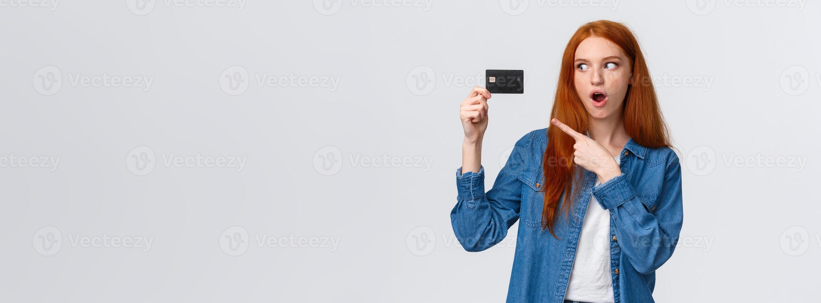 Discounts, shopping and banking concept. Waist-up impressed, thrilled cute redhead girl in denim shirt holding credit card, pointing at it with fascinated impressed eyes, standing white background photo