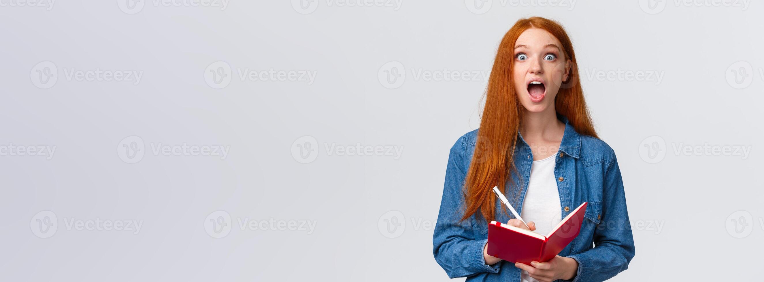 Amused and excited, astonished redhead girl fascinated with amazing lecture giving speech, writing down useful notes, holding notebook and staring thrilled camera, white background photo