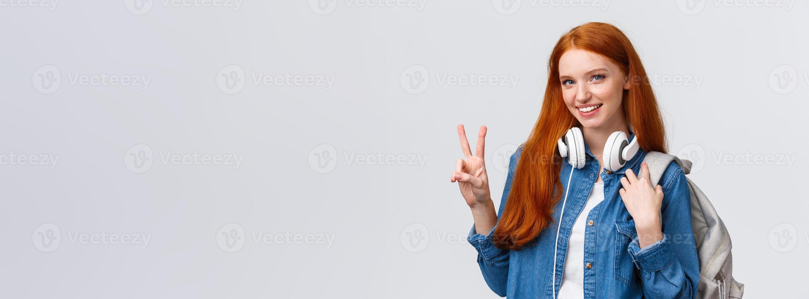 Lovely charismatic caucasian redhead girl with backpack, headphones over neck, showing peace sign and smiling delighted and joyful camera, standing white background, heading part-time job photo